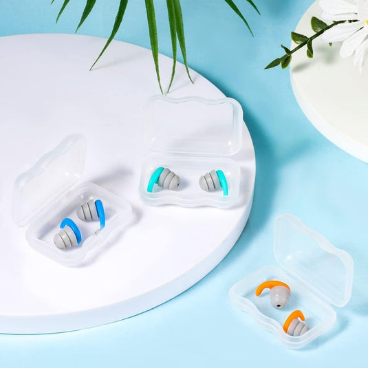 Drie paar grijze siliconen zwemoordopjes van Ervaar, met een waterdicht design met blauwe, turquoise en oranje lusjes, zitten in doorzichtige plastic doosjes op een wit en blauw oppervlak, omringd door groene bladeren en een witte bloem.