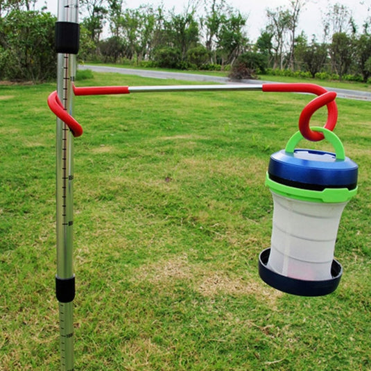 Het Avontuurlijk licht: 3x outdoor multifunctionele campinglamp haak hangt aan een rode haak aan een metalen paal met gras, bomen en een pad op de achtergrond - ideaal voor buitenavonturen.