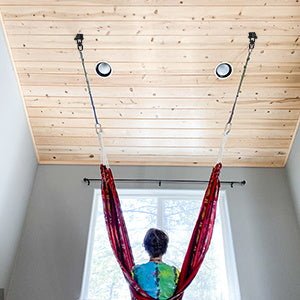 In een rode hangmat binnenshuis geniet men van het zonlicht dat door het houten plafond filtert, dankzij De ultieme 360° draaibare plafondhaak voor maximale veelzijdigheid en veiligheid, die een veilig en panoramisch uitzicht op de rustige omgeving mogelijk maakt.