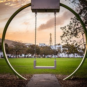 Laad afbeelding in Galerijviewer, Een moderne, ronde schommel van roestvrij staal staat leeg in het park, omlijst door bomen, met een grasveld en stadsgebouwen op de achtergrond, onder een oranje lucht.
