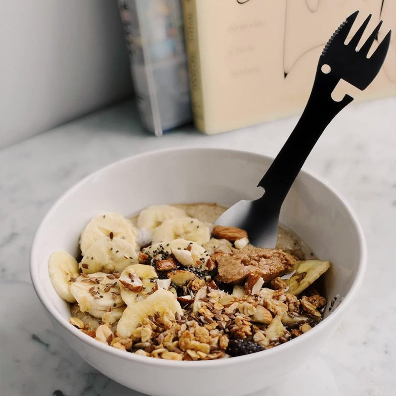 Laad afbeelding in Galerijviewer, Een kom havermout belegd met gesneden bananen, muesli, gehakte noten en zaden bevat de De ultieme multifunctionele campinglepel: Jouw beste vriend voor elk avontuur erin. Op de achtergrond zijn boeken zichtbaar.

