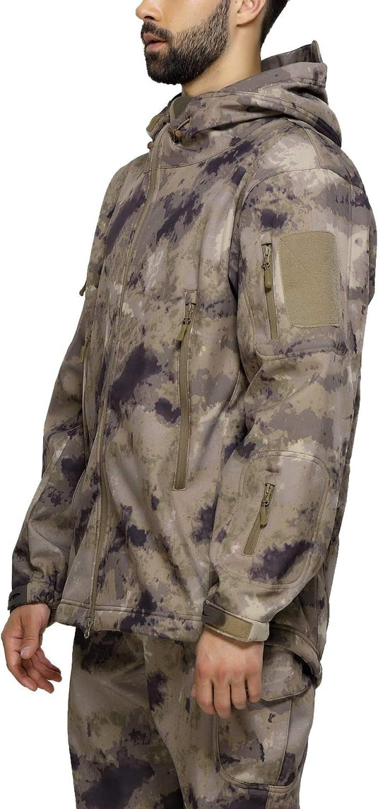 Een persoon in een camouflage "Camouflage tactische heren softshell jas: de perfecte metgezel voor je outdoor avonturen" en broek staat zijwaarts met ontspannen handen. De boksjas heeft meerdere zakken met ritssluiting en een capuchon voor extra functionaliteit.