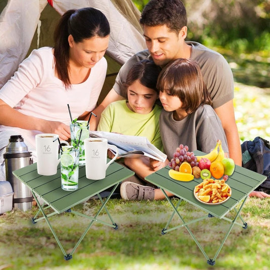 Een gezin geniet van een boek bij hun tent, zittend op het gras. Vooraan staan twee opvouwbare tafels met drankjes, fruit en snacks te midden van zonnig groen. Hun ultralichte draagbare kampeertafel van Ontdek heeft een antislip voetdopontwerp voor extra stabiliteit.
