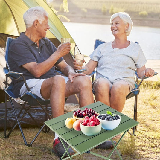 Een ouder echtpaar zit in kampeerstoelen en deelt een toost met een drankje. Voor hen staat onze "Ontdek de ultieme kampeerervaring met onze draagbare campingtafel!", beladen met schalen vol kersen, bessen en perziken. Een tent en waterpartij op de achtergrond versterken de rust en charme van het tafereel.