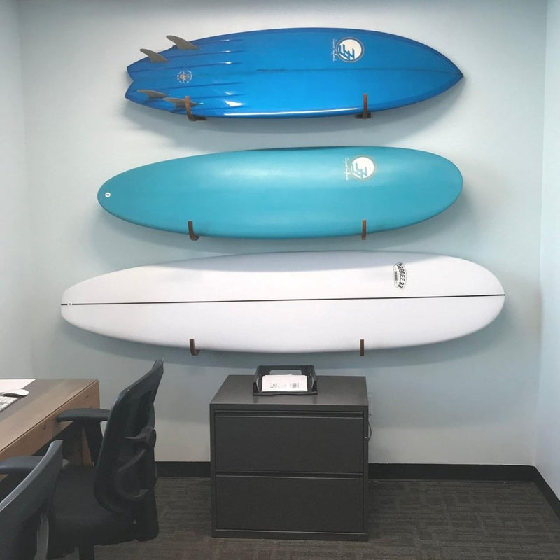 Laad afbeelding in Galerijviewer, Drie surfplanken in blauw en wit, elegant gemonteerd met de tekst &quot;Jouw surfplank verdient een stijlvolle display! 🏄‍♂️&quot;, zweven boven een zwarte archiefkast in een kantoorruimte met een bureau en een stoel.
