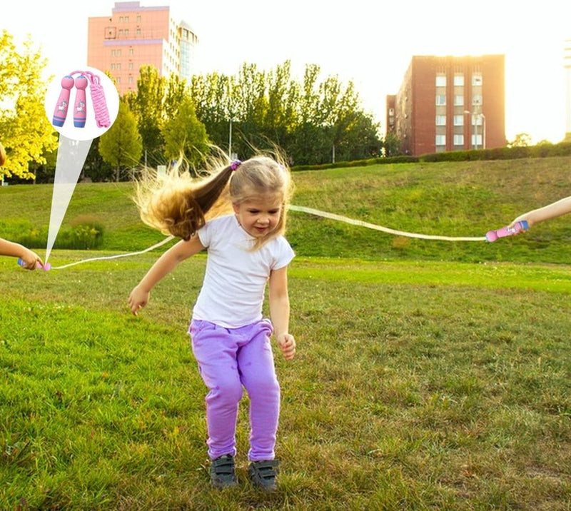 Laad afbeelding in Galerijviewer, Een kind met lang haar springt touwtjespringen op het gras, vastgehouden door twee mensen, terwijl gebouwen en bomen het tafereel omlijsten. Met behulp van &quot;Het perfecte kinderspringtouw voor eindeloos speelplezier!&quot; ondersteunt deze speelse activiteit de lichamelijke ontwikkeling in een buitenomgeving.
