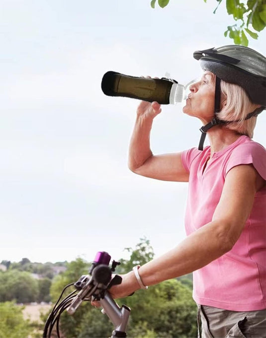 Een oudere vrouw in een roze shirt en helm drinkt uit de ultralichte herbruikbare siliconen sportfles, haar perfecte hydratatiepartner, terwijl ze een fiets vasthoudt tegen een achtergrond van groen gebladerte en een heldere lucht.