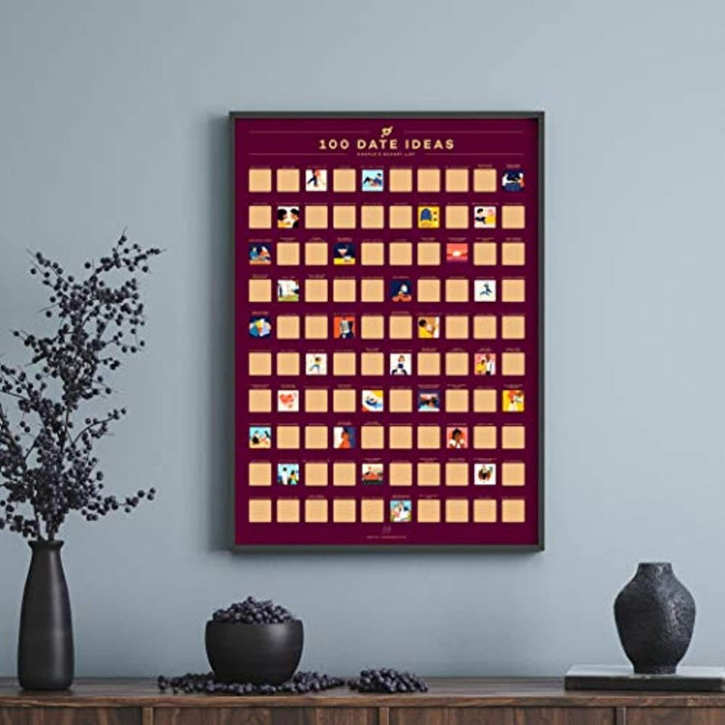 Laad afbeelding in Galerijviewer, De poster met de titel &quot;Ontdek 100 locatie afspraakjes met de romantische krasposter!&quot; hangt aan een grijze muur, waardoor het een perfect Valentijnscadeau is. Vlakbij staan een vaas met takken en een schaaltje op een donkere tafel, wat de romantische sfeer versterkt.
