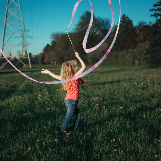 Een kind met lang haar draagt een rood shirt en draait sierlijk een roze lint in een grasveld met bomen en verre elektriciteitsleidingen, met behulp van Dansbanden voor kinderen: De ultieme manier om je dansroutines naar een hoger niveau te tillen.