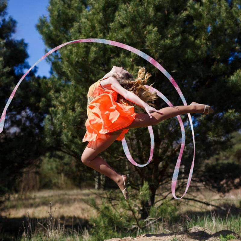 Laad afbeelding in Galerijviewer, In een hemelse gratie voert een danseres in een lichte oranje polyester jurk met Dansbanden voor kinderen een adembenemende sprong uit tegen een serene achtergrond van bomen en gras, die doet denken aan ritmische gymnastiek.
