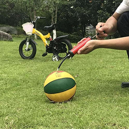 Met het symbool "Geniet overal van je spel met de draagbare ballenpomp" op gras blaast een persoon een geel-groene basketbal op, terwijl een klein fietsje met een wit mandje charme aan het tafereel toevoegt.