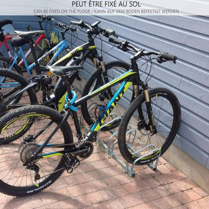 Laad afbeelding in Galerijviewer, Twee mountainbikes staan geparkeerd in een fietsenrek op een stenen terras bij een blauwe muur. Het &quot;Fietsenrek voor 3 of 4 fietsen&quot; is gemaakt van gegalvaniseerd staal, kan aan de vloer worden bevestigd en is voorzien van meertalige bewegwijzering voor efficiënte ruimtebesparing.
