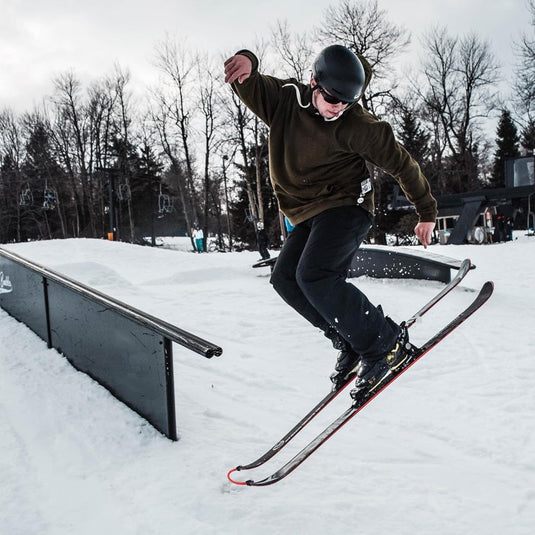 Tussen bladerloze bomen op een besneeuwde helling voert een skiër met helm en bril vakkundig een truc uit in de lucht bij een rail. Met behulp van de "Leer je kinderen skiën met deze skitipconnector" kunnen kinderen leren veilig te skiën met vertrouwen en flair.