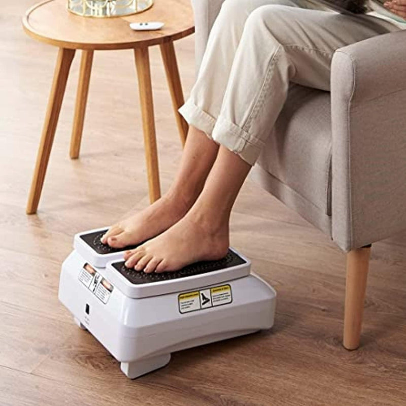 Laad afbeelding in Galerijviewer, Een persoon ontspant op een bank, met zijn voeten op de Beentrainer pro, een wit voetmassage-apparaat met dubbele functie en beentrainer, naast een houten tafel op de vloer.
