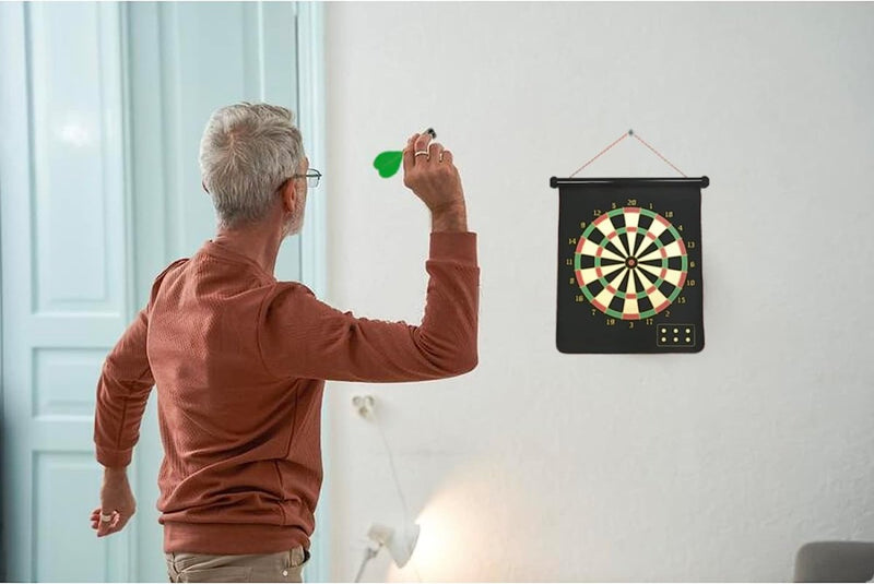 Laad afbeelding in Galerijviewer, Een oudere volwassene met grijs haar en een bril gooit een pijltje naar het Dubbelzijdige magnetische dartbord dat binnenshuis aan een witte muur hangt, zodat het veilig is voor kinderen.
