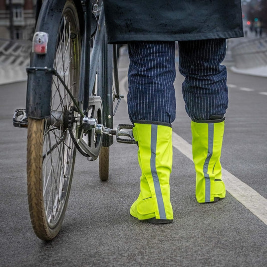 Een persoon staat zelfverzekerd naast een fiets op de verharde weg, met felgele reflecterende regenoverschoenen voor fietsers: blijf droog en stijlvol, die veiligheid en bescherming belichamen.