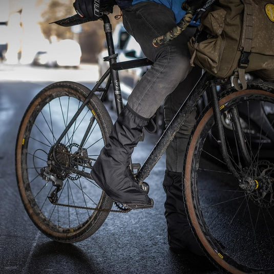Een persoon op een zwarte fiets trapt zelfverzekerd in regenoverschoenen voor fietsers, gekleed in een donkere spijkerbroek. Het blauwe jack biedt extra bescherming en een groene tas is stevig aan de fiets bevestigd.