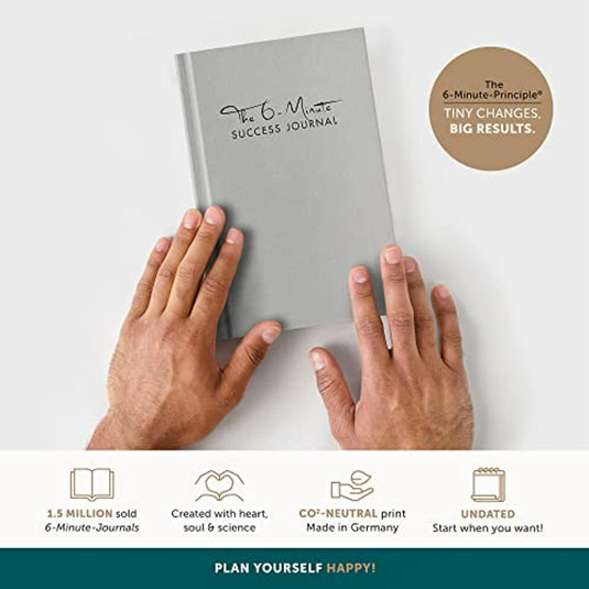 Twee handen die "Het 6-Minute Success Journal" vasthouden benadrukken mindfulness. Belangrijke kenmerken zijn CO₂-neutraal printen en een ongedateerd formaat, allemaal ontworpen om de productiviteit in slechts zes minuten per dag te verbeteren.