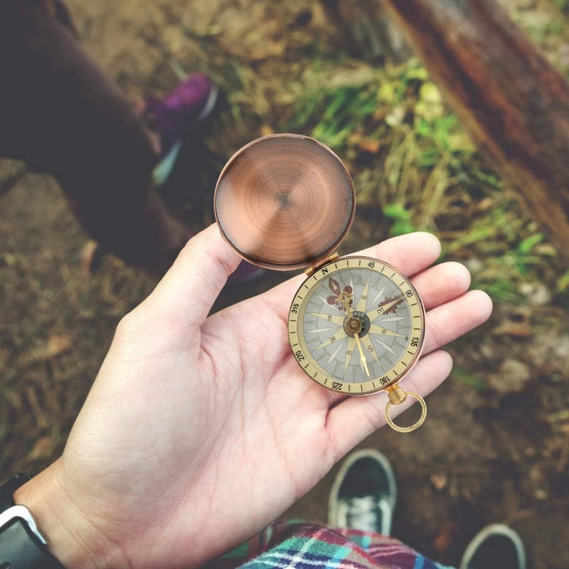 Laad afbeelding in Galerijviewer, Een hand houdt de Vintage koperkompas vast: een klassiek hulpmiddel voor outdoorliefhebbers, die kardinale richtingen onthult, met het koperen deksel ernaast. Het robuuste, compacte ontwerp rust tegen een met gras begroeide achtergrond en een wazige voet.
