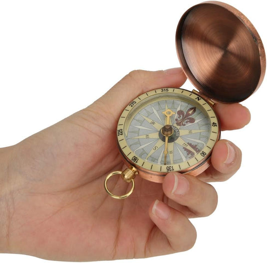 Een hand houdt een open Vintage koperkompas vast: een klassiek hulpmiddel voor outdoorliefhebbers, met een decoratief gezicht en lus. Deze compacte en lichtgewicht schat is zowel praktisch als robuust voor elk avontuur.