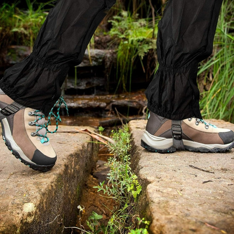Laad afbeelding in Galerijviewer, Een persoon draagt beige en zwarte wandelschoenen met blauwgroene veters en het opschrift &quot;Gaiters: de ultieme bescherming voor al je buitenactiviteiten&quot; en steekt een kleine rotsachtige kloof over, omgeven door weelderig groen.
