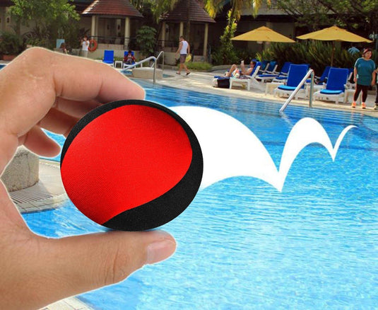 Een hand die een Waterballen vasthoudt: een waterpret voor het hele gezin, rood en zwart gekleurd, boven een zwembad met een geanimeerd wit spatpad op de achtergrond, perfect voor kinderen om van te genieten op een zonnige dag.